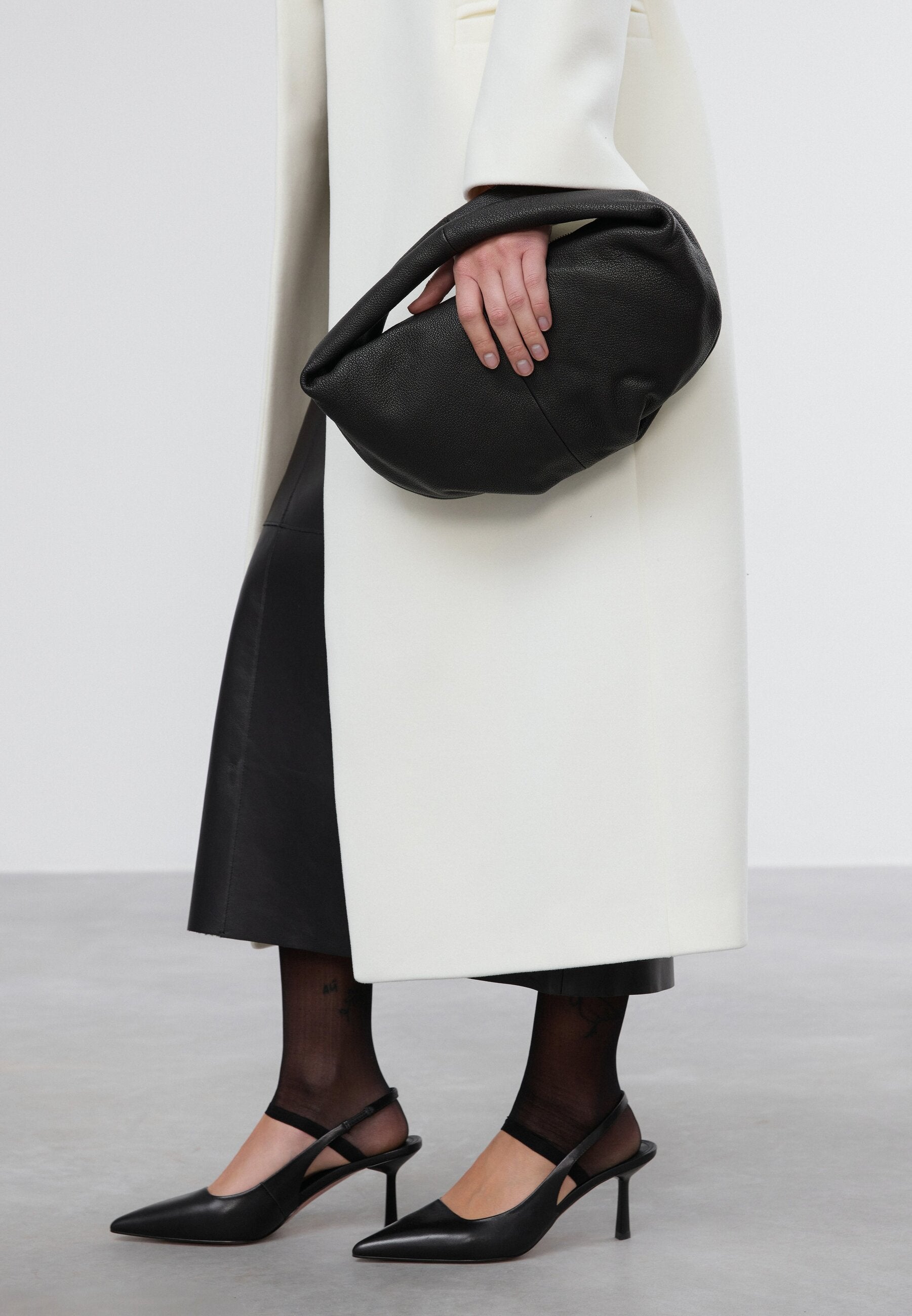 Model im weißen Mantel hält die schwarze Tasche lässig unter dem Arm geklemmt als Oversize-Clutch