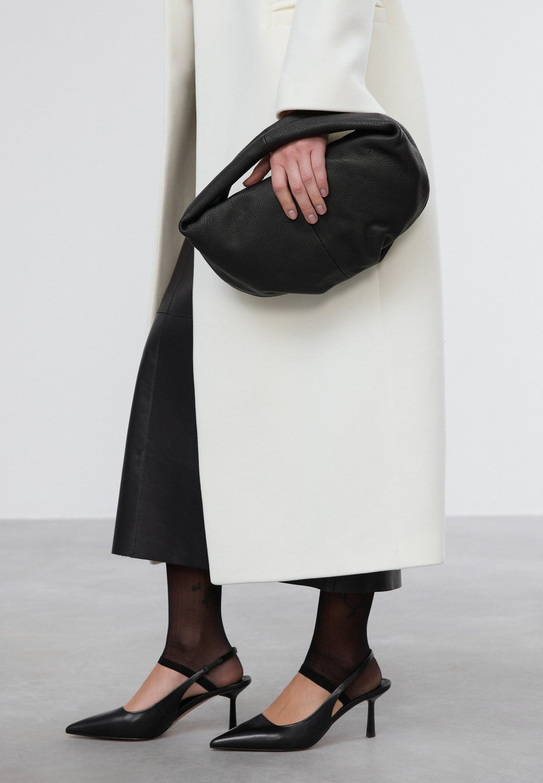 Model im weißen Mantel hält die schwarze Tasche lässig unter dem Arm geklemmt als Oversize-Clutch