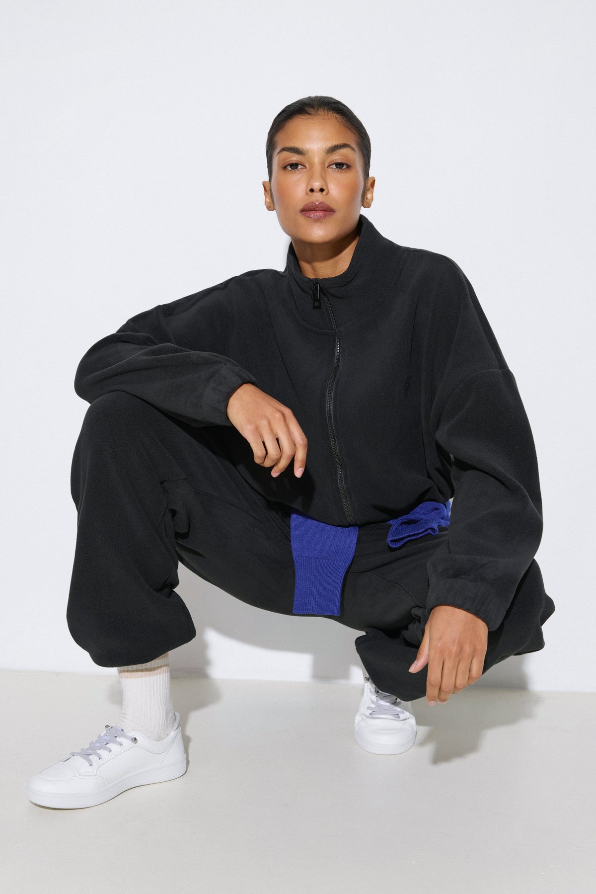 Kauerndes Model im schwarzen Damen-Fleece-Set von Estro im Loose Fit, gestylt mit einem blauen Pullover um die Taille