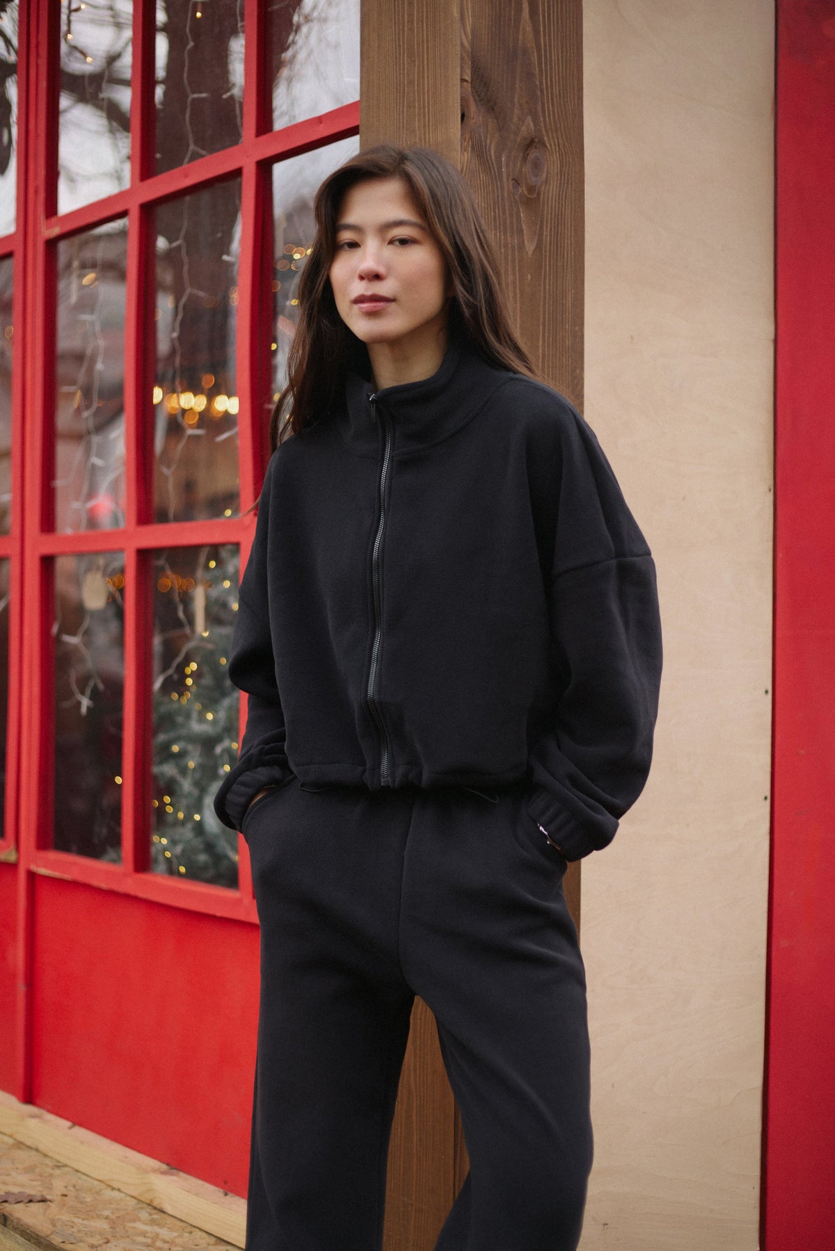 Outdoor-Lifestyle-Aufnahme des Models im schwarzen Fleece-Set vor einer rustikalen roten Fensterfront