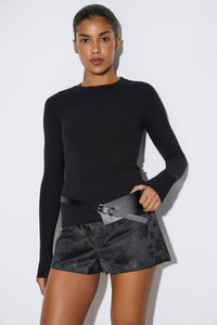Studio-Aufnahme 3/4-Profil: Model im schwarzen gerippten Damen-Longsleeve von Estro Club, kombiniert mit gemusterten Shorts, Gürtel und hohen Stiefeln