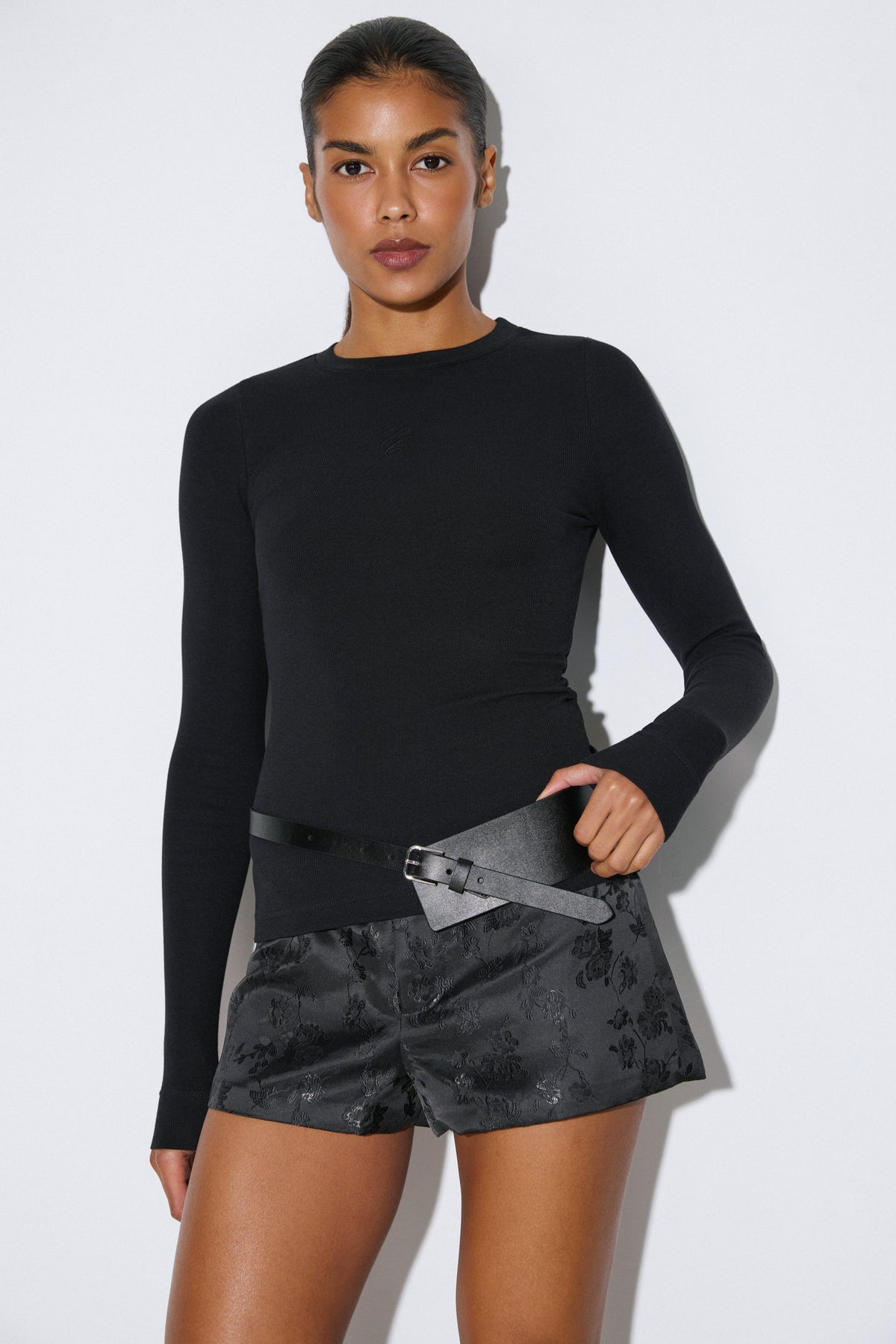 Studio-Aufnahme 3/4-Profil: Model im schwarzen gerippten Damen-Longsleeve von Estro Club, kombiniert mit gemusterten Shorts, Gürtel und hohen Stiefeln